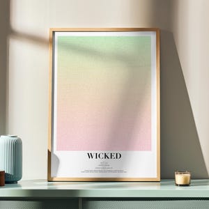 Puede incluir: Una impresión artística enmarcada con un degradado de verde, amarillo y rosa. La palabra "WICKED" está impresa en negro en la parte inferior. Un jarrón azul, una caja de madera y una vela encendida están sobre una superficie verde menta.