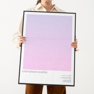 Op de afbeelding: Een ingelijste poster met een gradiëntachtergrond van paarse en roze tinten. De poster heeft de tekst "K-POP DEMON HUNTERS" onderaan, met extra tekst eronder. De lijst is zwart.