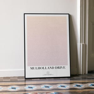Puede incluir: Un póster enmarcado con un degradado de rosa y melocotón. El título "Mulholland Drive" se muestra en negrita. El póster está apoyado contra una pared blanca y un suelo estampado.