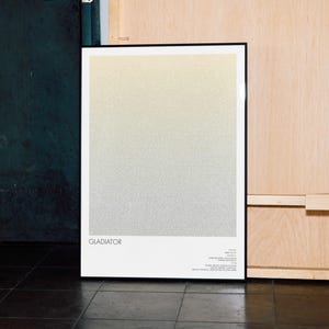 May include: A framed poster with a gradient design, transitioning from light yellow to gray. The poster has a black frame and the word "GLADIATOR" is printed at the bottom. Additional text is visible below the title.