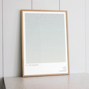 May include: A framed poster with a light wood frame. The artwork features a gradient of light blue and gray. The text "FI: THE MOVIE" is at the bottom left, with additional text on the bottom right. The poster is leaning against a white wall.