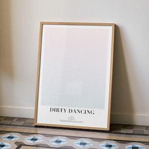 May include: A framed poster with the text "DIRTY DANCING" in black. The poster has a gradient background of pink, white, and light blue. The frame is made of light brown wood. The poster is leaning against a white wall.