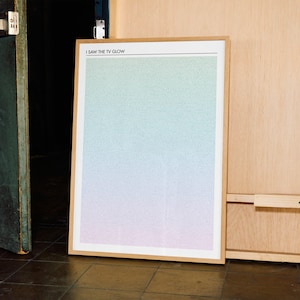 May include: A framed art print with a gradient background of green, blue, and pink hues. The print is in a light wood frame and features the text "I SAW THE TV GLOW" at the top. The artwork is leaning against a wooden wall.