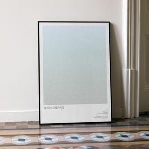 Könnte beinhalten: Ein gerahmter Kunstdruck mit minimalistischem Design. Das Kunstwerk zeigt einen Farbverlauf aus Blau- und Grautönen mit den Worten "TRAIN DREAMS" am unteren Rand. Der Druck lehnt an einer weißen Wand neben einer weißen Tür.