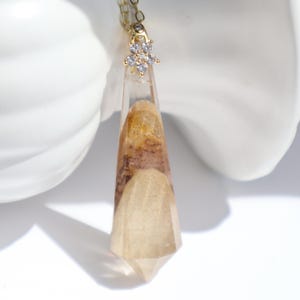 Può includere: Una collana color oro con un ciondolo geometrico trasparente. Il ciondolo contiene un mix di materiale organico marrone e beige. La collana presenta un piccolo abbellimento a forma di fiore con pietre trasparenti.