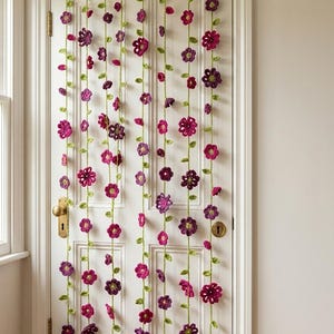 Puede incluir: Una puerta blanca adornada con guirnaldas de flores de ganchillo. Las guirnaldas presentan flores rosas y moradas con tallos y hojas verdes, creando un efecto de cortina decorativa. La puerta tiene un pomo y una cerradura de latón.