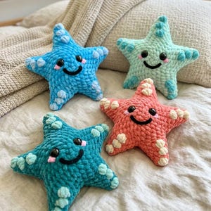 Puede incluir: Cuatro estrellas de mar de ganchillo hechas a mano en tonos de azul, verde azulado y coral, cada una con una cara sonriente y detalles blancos. Estos juguetes de peluche están colocados sobre una cama, mostrando un tema náutico caprichoso.