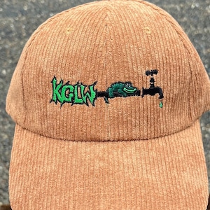 May include: A corduroy baseball cap in a warm brown color. The cap features an embroidered design with the letters "KGLW" in green, along with a green frog and a black faucet with a water drop.