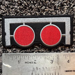 May include: Embroidered patch featuring two red circles with white outlines on a black background. The patch has a rectangular shape with a silver border and measures approximately 4 inches in length.