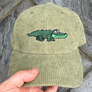May include: A green corduroy baseball cap with an embroidered alligator design. The alligator is green with black eyes and a white mouth.