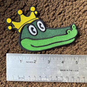May include: Green embroidered alligator patch with a yellow crown. The alligator is smiling with large white eyes. The patch is approximately 2 inches wide.