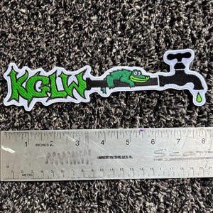May include: Embroidered patch featuring the text "KGLW" in green graffiti-style lettering. The design includes a green alligator resting on a black faucet with a green water droplet. The patch is approximately 7 inches long.