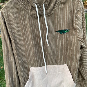 May include: A brown corduroy hoodie with a white drawstring and a small green alligator embroidered on the front. The hoodie has a large beige corduroy pocket on the front with a small black patch that says "POINT 7 THE GAME OF LIFE"