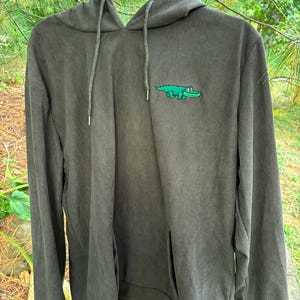 May include: Olive green corduroy hoodie with a kangaroo pocket and drawstring hood. A green alligator graphic is embroidered on the chest. The hoodie is hanging on a black hanger against a backdrop of green foliage.