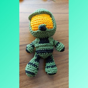 May include: Green crocheted plush toy of a video game character wearing a helmet with a yellow visor. The toy has black accents on the body and helmet.