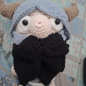 May include: A handmade crochet doll in the shape of a Viking. The doll has a blue helmet, tan horns, a black beard, and a pink nose. The eyes are black. The doll is made of yarn and has a whimsical design.