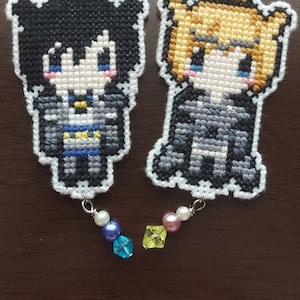 May include: Two embroidered keychains featuring cartoon characters. The keychain on the left features a black-haired character with a blue and yellow outfit. The keychain on the right features a blonde character with a gray and white outfit. Both keychains have a silver chain and a white pearl and crystal bead charm.