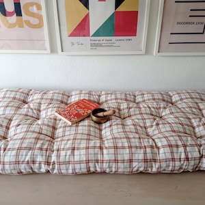 May include: A large, tufted floor cushion with a red and white plaid pattern. A red book and a small brass singing bowl with a wooden striker rest on the cushion. Decorative framed art hangs on the wall above.