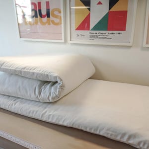 Può includere: Un set di materasso e cuscino bianchi piegati su una superficie marrone chiaro. La biancheria da letto è ben disposta, con il cuscino piegato sopra il materasso. Lo sfondo presenta opere d'arte incorniciate.