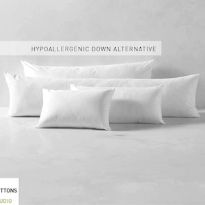 Könnte beinhalten: Vier weiße hypoallergene Daunen-Alternative-Kissen in verschiedenen Größen. Die Kissen sind pyramidenförmig auf einer weißen Oberfläche angeordnet. Der Text "HYPOALLERGENIC DOWN ALTERNATIVE" wird über den Kissen angezeigt.