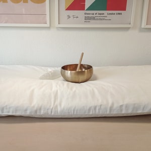 Può includere: Un cuscino da meditazione bianco con una ciotola cantate dorata e un battitore di legno in cima. Il cuscino è su una superficie di legno.