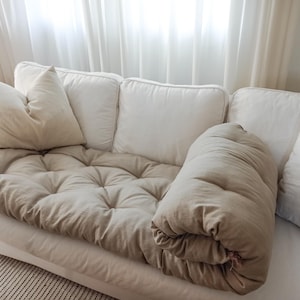 Könnte beinhalten: Ein weißes Sofa mit einem getufteten, beigefarbenen Kissen und mehreren weißen Kissen. Eine zusammengerollte beige Decke liegt auf dem Sofa. Die Szene wird von natürlichem Licht von einem durchsichtigen Vorhangfenster beleuchtet.