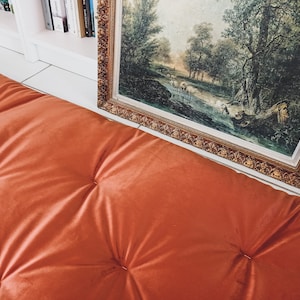 May include: A close-up of a burnt orange velvet cushion with button tufting. The cushion is in front of a framed landscape painting with a gold frame.