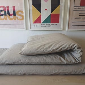 May include: Beige linen bedding set, comprising a folded duvet and pillows, arranged on a wooden surface. The background displays framed art prints with geometric designs and text. The image highlights the fabric's texture and is well-lit.