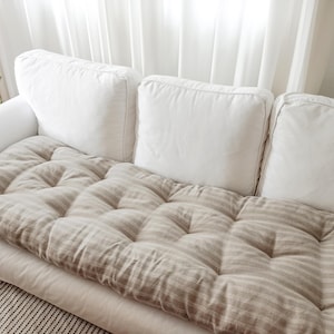 May include: A white sofa with three white back cushions and a tufted, striped cushion. The striped cushion is beige and has a series of evenly spaced, vertical stripes. The sofa is in front of a white curtain.