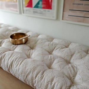 Könnte beinhalten: Ein getuftetes Bankkissen mit cremefarbenem und beigem Muster, gekrönt von einer kleinen Messingschale und einem Holzschlägel. Das Kissen liegt auf einer Holzoberfläche, im Hintergrund gerahmte Kunstwerke.