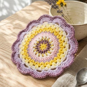 May include: A handmade, colorful crochet coaster with a scalloped edge. The coaster features concentric circles of yellow, pink, and purple. It sits on a wooden surface, next to a spoon and a linen napkin.