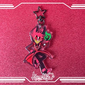 May include: A clear acrylic keychain featuring a cartoon character in a red suit with black pants, holding a green flame. The keychain has a star-shaped clasp and a floral logo at the bottom. The background is a solid red.