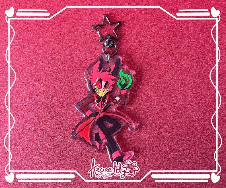 May include: A clear acrylic keychain featuring a cartoon character in a red suit with black pants, holding a green flame. The keychain has a star-shaped clasp and a floral logo at the bottom. The background is a solid red.