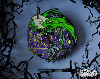 May include: A green and purple enamel pin featuring a cartoon character with a witch's hat riding a green monster with a skeletal body. The pin has a glittery background with stars and a purple circle with a symbol in the center.