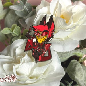 May include: A red and black enamel pin featuring a cartoon character with red horns, a red suit, and a wide grin. The character is holding a microphone in one hand and has a devilish expression.