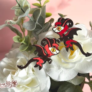 May include: Two red and black enamel pins of a cartoon dragon with wings and horns. The pins are on a white flower background.