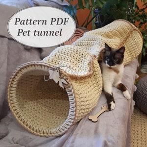 May include: A beige crocheted pet tunnel with a brown trim. A cat is partially inside the tunnel, and a wooden bone toy is on the floor. The text "Pattern PDF Pet tunnel" is in a white circle on the top left of the image.