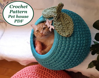 May include: A teal blue crocheted pet house shaped like an apple with a green crocheted leaf on top. A ginger cat is inside the house. The house has a small opening for the cat to enter and exit. The house is sitting on a pink crocheted cushion.