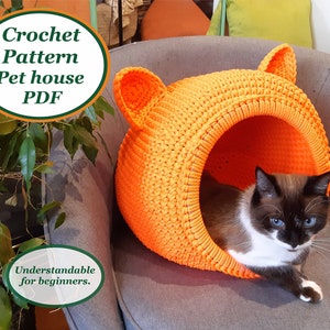 Pode incluir: Uma casa para animais de estimação de cor laranja feita de crochê com orelhas de gato, projetada para animais de estimação pequenos. A casa está em uma cadeira cinza com um gato dentro. O texto "Crochet Pattern Pet house PDF Understandable for beginners." está na imagem.