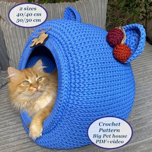 May include: A blue crocheted cat bed in the shape of a house with a cat inside. The bed has two red and orange pom-poms on the side. The text "Crochet Pattern Big Pet house PDF+video" is on the bottom of the image.