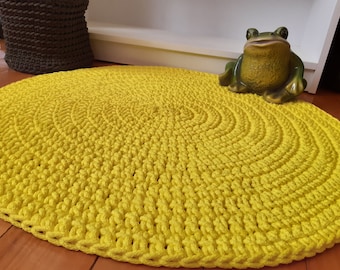 May include: A bright yellow, oval-shaped, crocheted rug. The rug has a spiral design and is made of thick yarn. A ceramic frog figurine is on the right side of the rug. A brown woven basket is in the background.