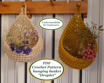 May include: Two crocheted hanging baskets, one beige and one mustard yellow, with dried flowers inside. The baskets are hanging from a wooden rack against a striped wall. The text "PDF Crochet Pattern hanging basket "Droplet"  Understandable for beginners." is below the baskets.