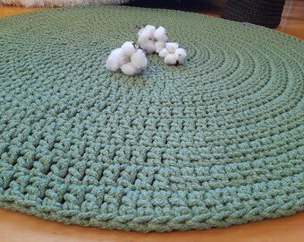 May include: A round, green crocheted rug with a textured surface. The rug is made of thick yarn and has a natural, earthy look.