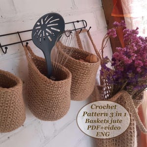 Puede incluir: Tres cestas de yute tejidas a crochet colgadas de un estante de metal negro. Las cestas son de color beige y tienen una textura tejida. Las cestas están etiquetadas "Crochet Pattern 3 in 1 Baskets Jute PDF+video ENG".
