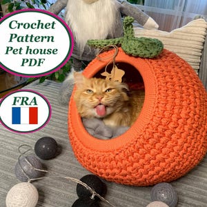 Puede incluir: Una casa para mascotas de ganchillo naranja con forma de fruta, con un tallo verde. Un gato jengibre está dentro de la casa, sacando la lengua. El texto "Crochet Pattern Pet house PDF" está en un círculo encima.