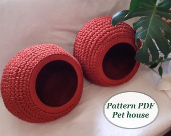 May include: Two red crocheted pet houses, shaped like a dome with a round opening. The houses are sitting on a white surface with a green plant in the background. The text "Pattern PDF Pet house" is written in white on a gray oval.