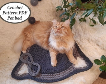 May include: A crocheted pet bed in a dark grey and light grey colour scheme. The bed is shaped like a cat's head with a large, grey, crocheted infinity symbol in the centre. A ginger cat is lying on the bed, looking up at the camera. The text "Crochet Pattern PDF Pet bed" is visible in the top left corner of the image.