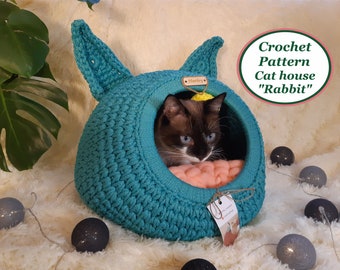 May include: A teal blue crocheted cat house shaped like a rabbit with a cat inside. The cat house has two floppy ears and a round opening. The cat is a Siamese with blue eyes and is lying on a pink blanket inside the house. The cat house is sitting on a white fluffy surface with gray string lights.