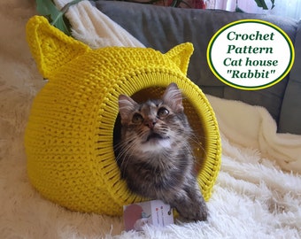 Könnte beinhalten: Ein gelbes Häkel-Katzenhaus in Form eines Kaninchens mit einer Katze darin. Das Katzenhaus befindet sich auf einer weißen, flauschigen Oberfläche. Crochet Pattern Cat house "Rabbit"