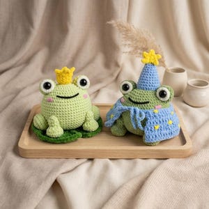 May include: Two handmade crochet frog figurines. One frog wears a gold crown, the other a blue party hat and shawl with yellow stars. Both are light green with large eyes and sit on a wooden tray.